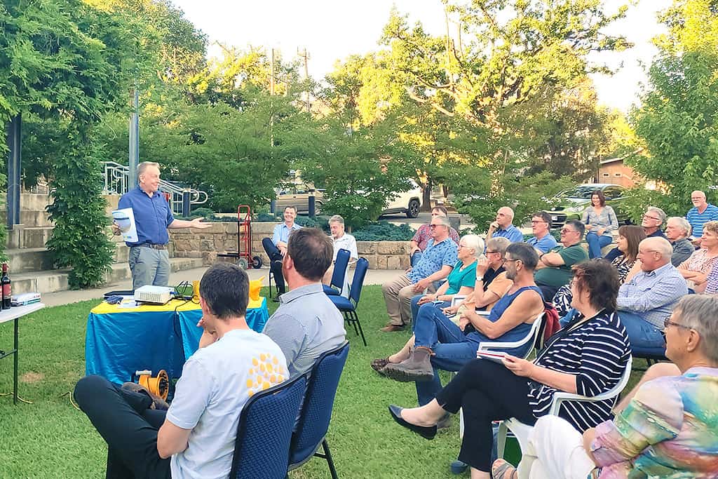 A man is talking to an audience of people on chairs on a lawn