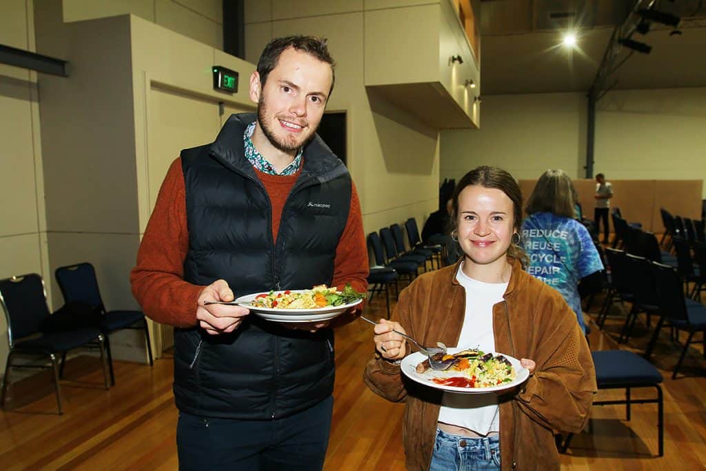 A man and a woman are standing with plates of food smiling at the camera