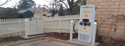 A white and cream narrow vertical EV charger unit with a yellow yak on it sits on the curb in front of a brick wall and next to a picket fence