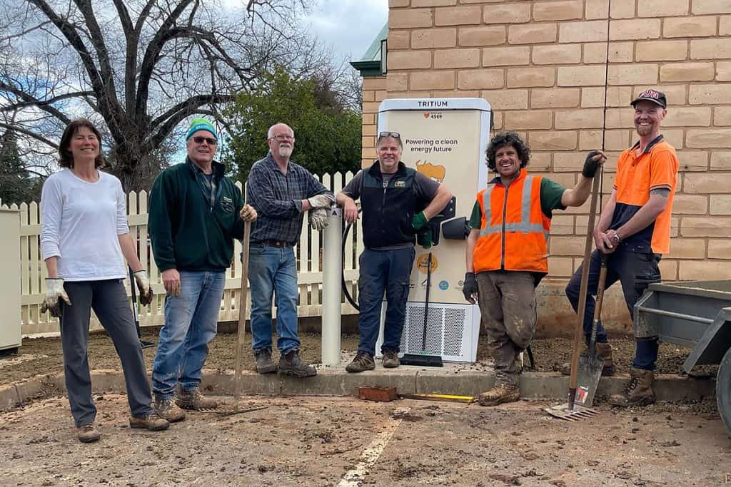 Six people with gloves, rakes, shovels and brooms stand in front of a cream and white EV charger unit on the edge of a carpark, looking towards and smiling at the camera.