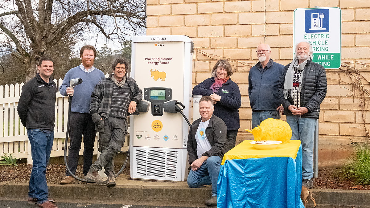 Seven people stand in front of a Tritium EV charger, on the edge of a car park, with a blue and yellow covered table and a yellow yak figurine in front of them.