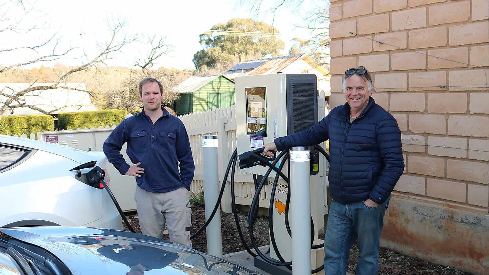 Two men stand on either side of an EV charger as it charges their car